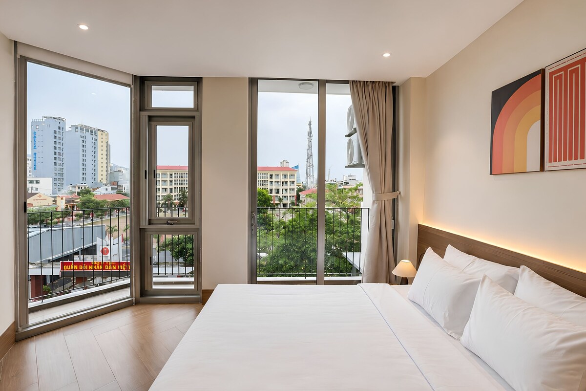 A hotel room with a large bed, a view of the city, and a balcony with a railing.