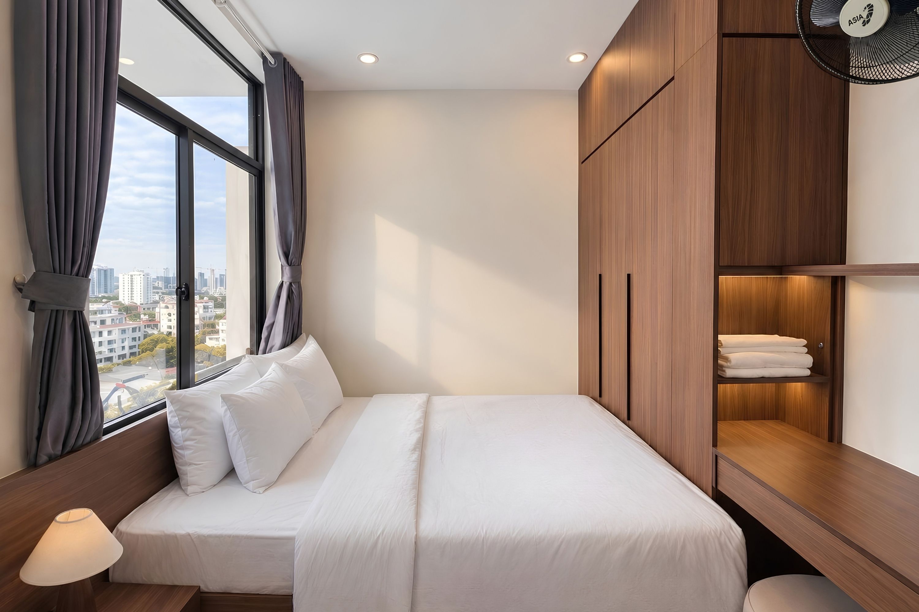 A hotel room with a bed, a nightstand, a lamp, a window with curtains, and a wardrobe.