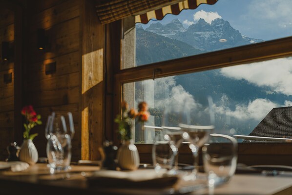 A table set with glasses and a vase of flowers, with a mountain view through a window.