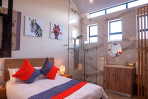 A modern bedroom with a bed, a wooden headboard, a nightstand, a bedside lamp, a glass shower enclosure, a marble wall, a wooden vanity with a mirror, and artwork on the wall.