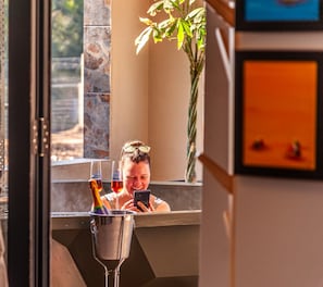 A person in a bathroom with a mirror, a plant, and a bottle of champagne. - Glass Castle 3 Bed Mountain View (Sir Lowry's Pass)