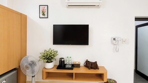A hotel room with a flat-screen TV mounted on the wall, a standing fan, a small shelf with a potted plant, a coffee maker, and a telephone. - Cat Ba Thai Long Hotel (Cat Hai)