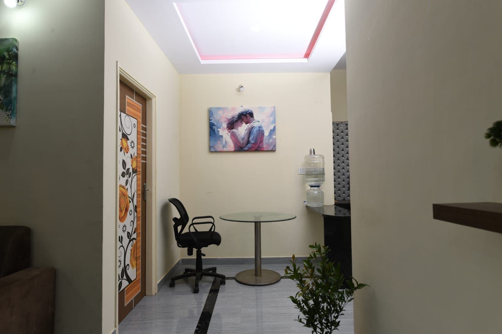 A hallway with a ceiling light, a painting, a water dispenser, a chair, and a small table.