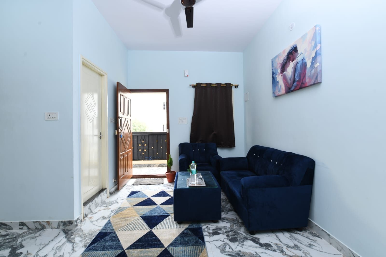 A living room with a blue sofa, a coffee table, a patterned rug, a painting on the wall, and a door leading to an outdoor area.