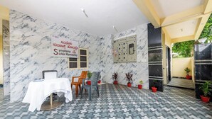 Habitación de hotel con pared de mármol, una mesa con mantel blanco y varias sillas. Hay plantas en macetas y un panel de control en la pared.