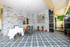 Habitación de hotel con pared de mármol, una mesa con mantel blanco y varias sillas. Hay plantas en macetas y un panel de control en la pared.