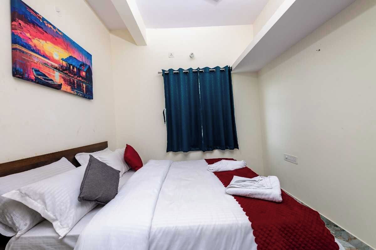 A hotel room with a bed, a red blanket, a painting on the wall, and a window with curtains.