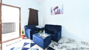 Una habitación con un sofá azul, una mesa de centro de vidrio, un cuadro en la pared y vistas a través de una puerta de cristal.