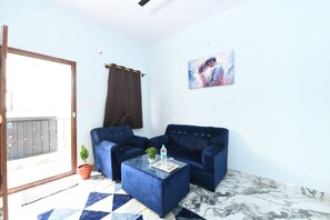 Una habitación con un sofá azul, una mesa de centro de vidrio, un cuadro en la pared y vistas a través de una puerta de cristal.