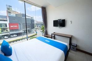 A hotel room with a large window, a bed with blue bedding, a small table, a TV on the wall, and a trash bin.