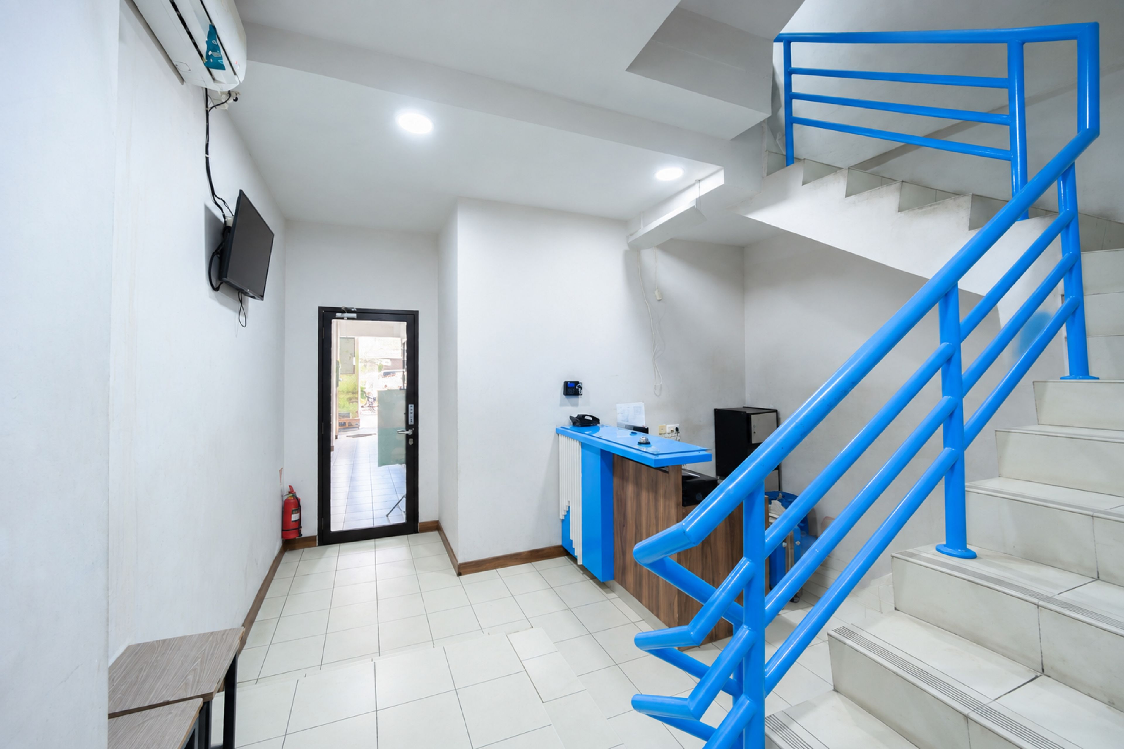 Un vestíbulo de hotel con un mostrador de recepción, una escalera con barandillas azules y un extintor cerca de la entrada.