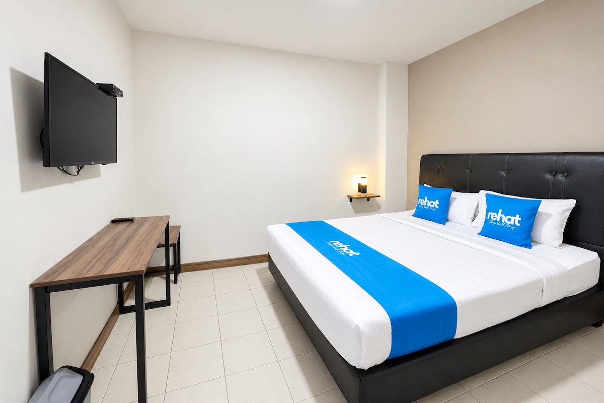 Habitación de hotel con cama, escritorio, silla, televisión de pantalla plana montada en la pared y mesita de noche con lámpara.