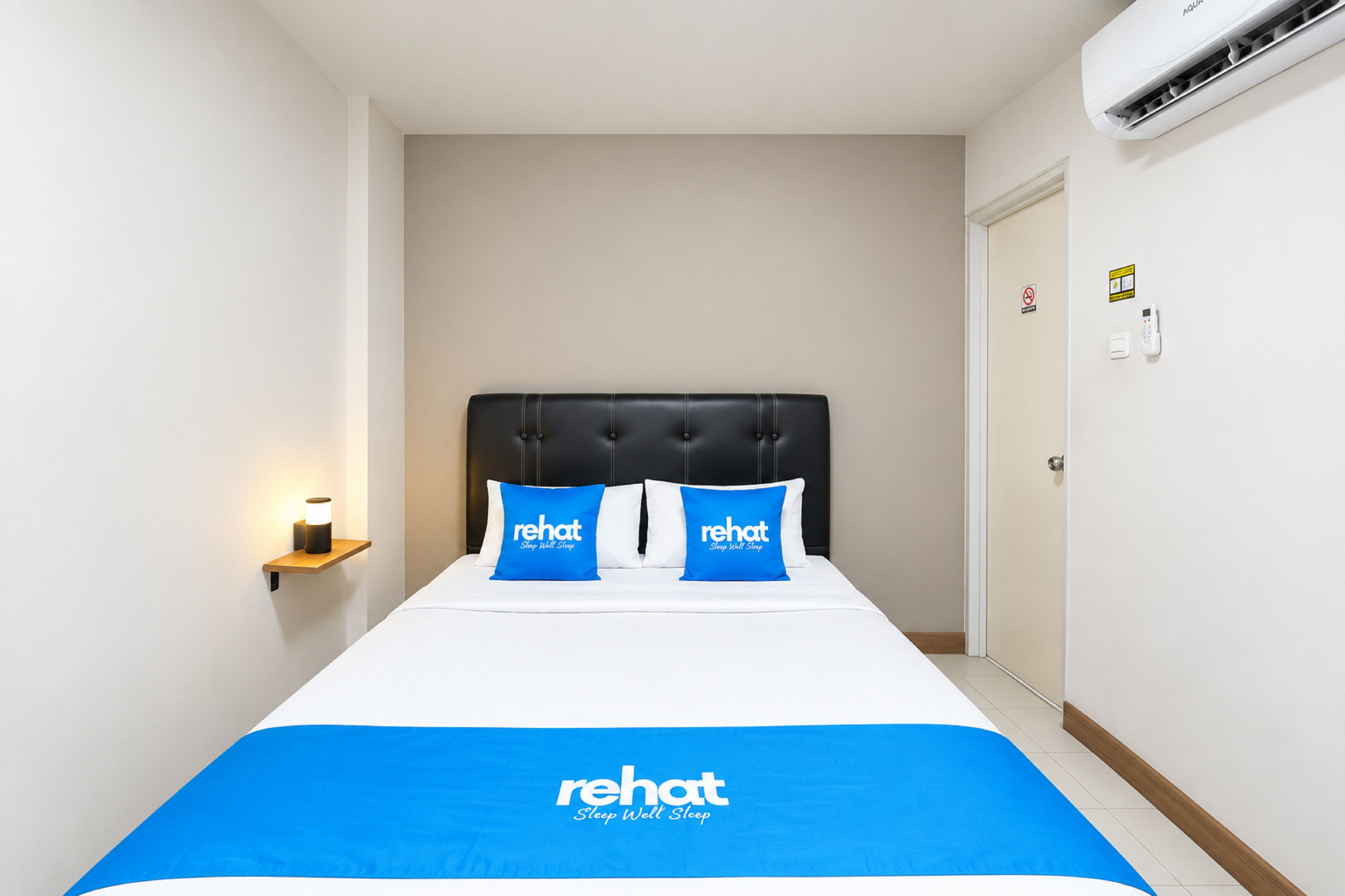 Una habitación de hotel con cama, dos almohadas con la marca 'rehat', una lámpara de noche y un aire acondicionado instalado en la pared.