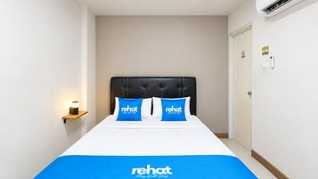 Una habitación de hotel con cama, dos almohadas con la marca 'rehat', una lámpara de noche y un aire acondicionado instalado en la pared.