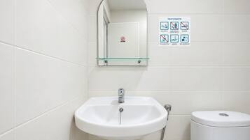 A bathroom with a white sink, a glass shelf above it, and a toilet. There is a sign with toilet etiquette guidelines on the wall.