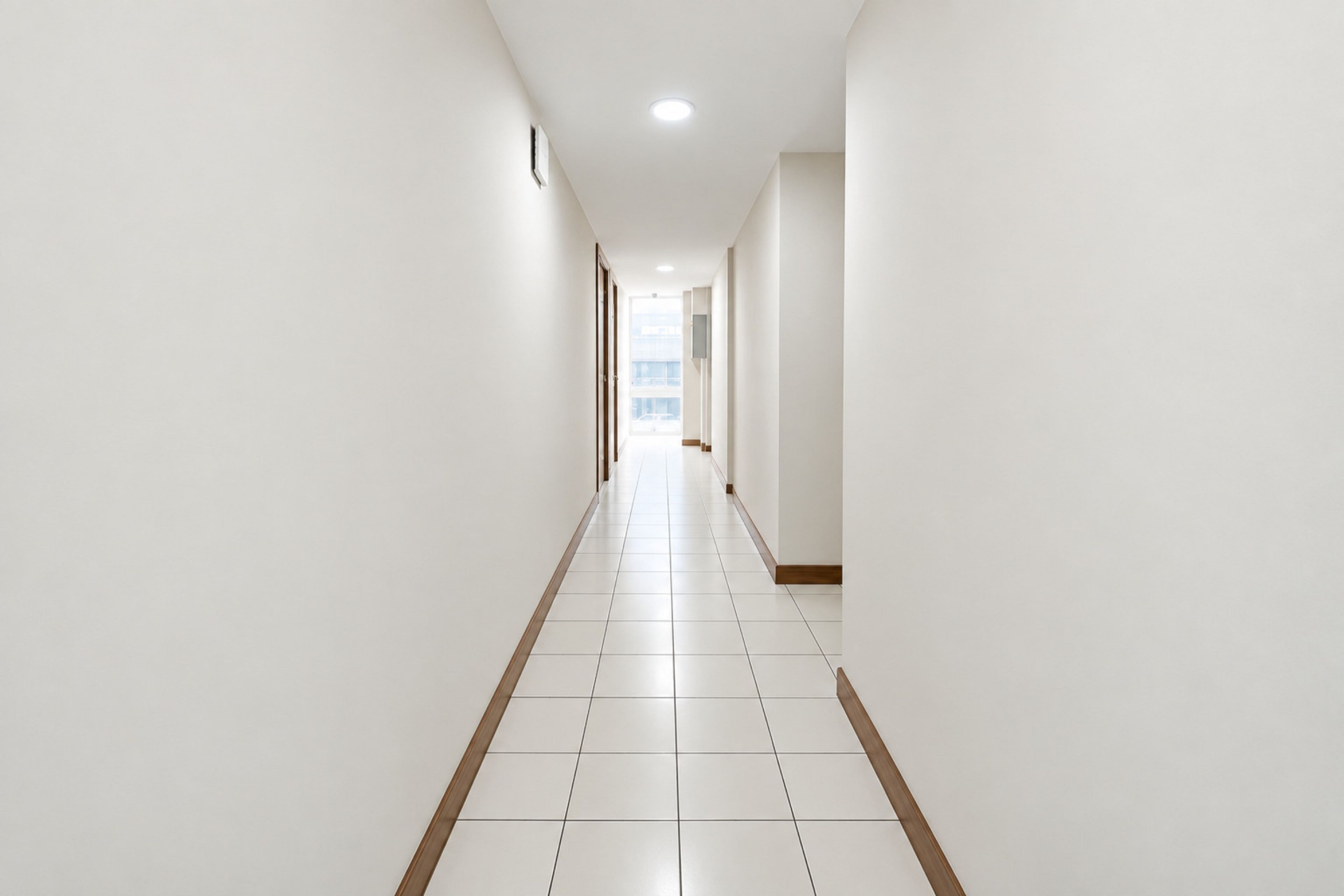 A long, narrow hallway with white walls and a tiled floor, leading to a bright area at the end.
