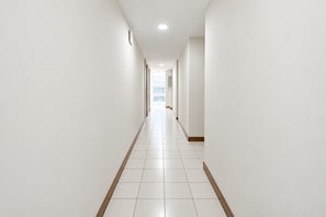 A long, narrow hallway with white walls and a tiled floor, leading to a bright area at the end.