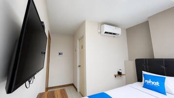 A hotel room with a flat-screen TV mounted on the wall, a bed with a blue pillow, and an air conditioning unit on the wall.
