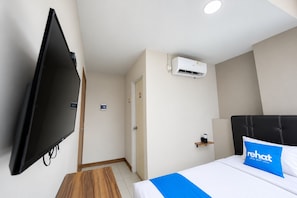 Una habitación de hotel con una televisión de pantalla plana montada en la pared, una cama con almohada azul y un equipo de aire acondicionado en la pared.
