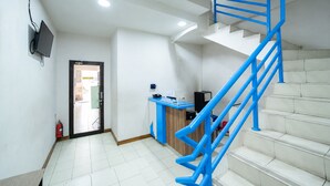 Escadaria com corrimãos azuis que leva a um andar superior, uma mesa de recepção e um extintor de incêndio no chão.