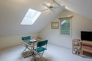 Une pièce avec un plafond en pente, un ventilateur de plafond, une table avec des chaises, une fenêtre et un téléviseur posé sur un meuble.