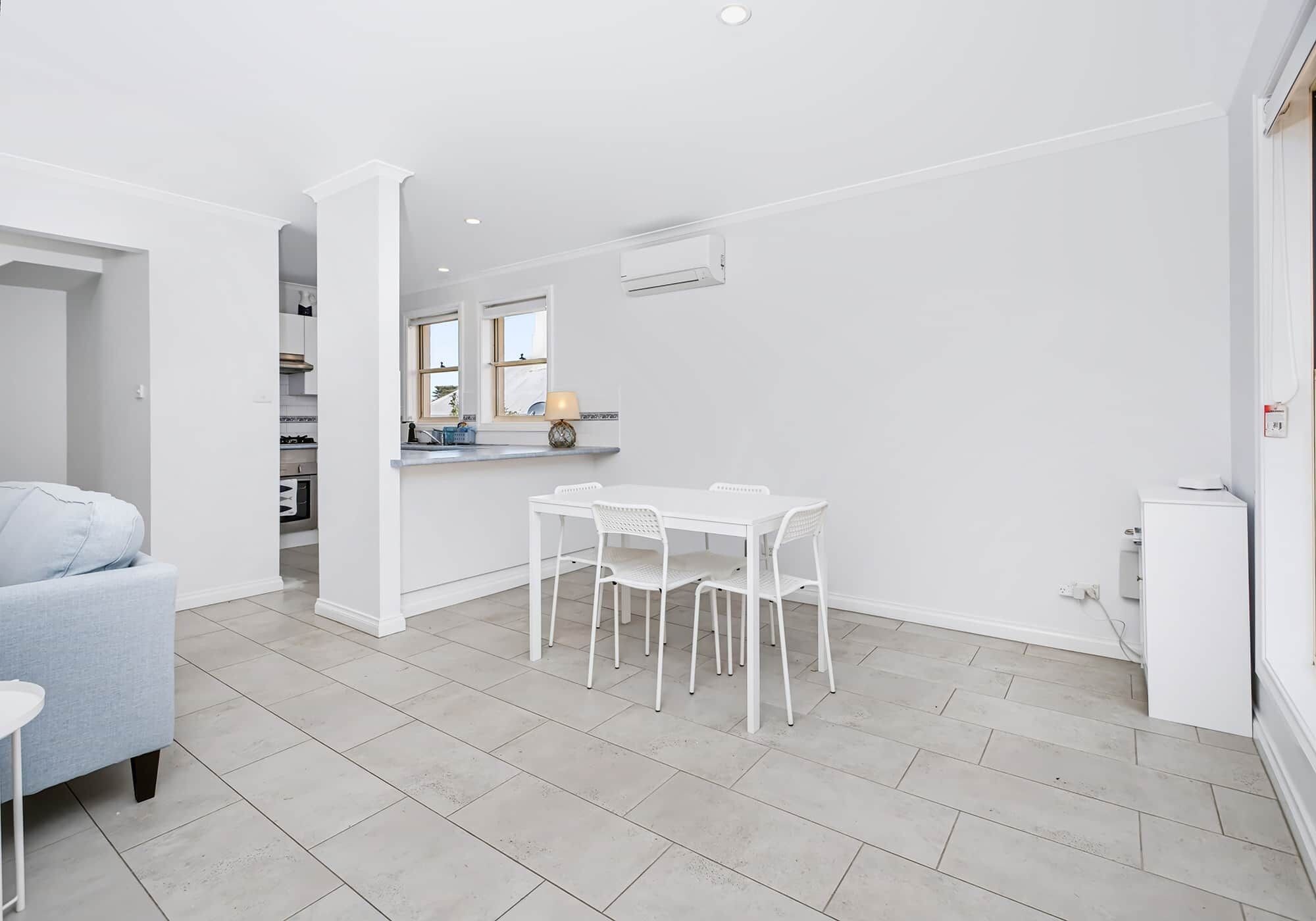 A modern kitchen with white cabinets, a dining area with white chairs, and a grey sofa.