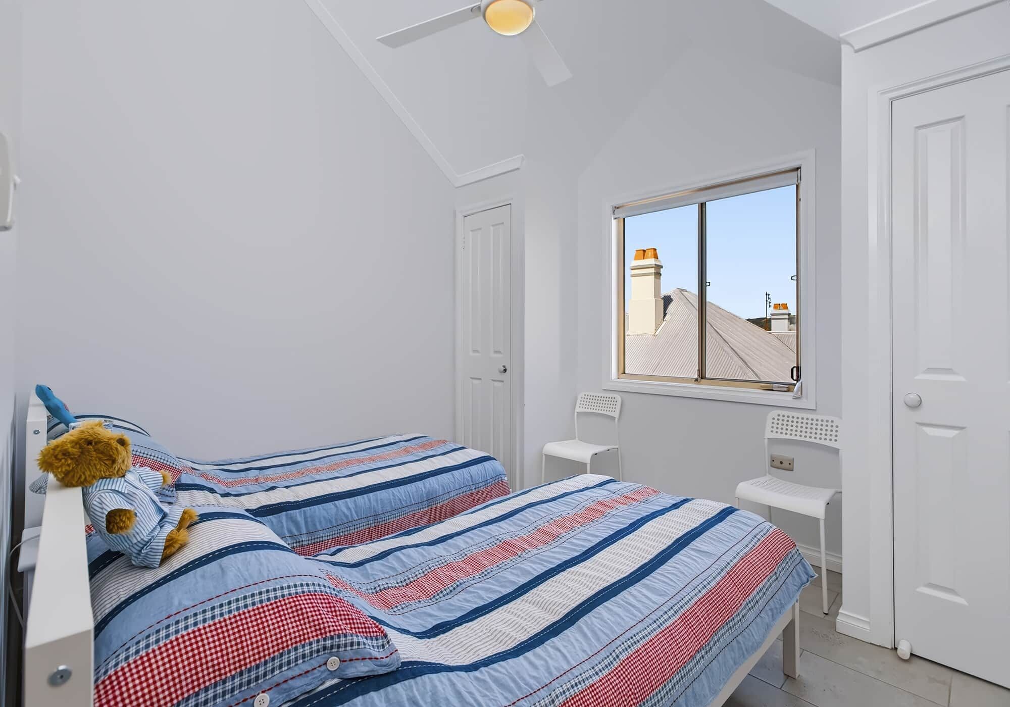 A bedroom with two beds, a window with a view of buildings, and a ceiling fan.
