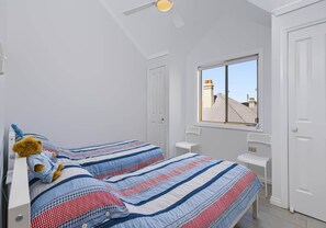 A bedroom with two beds, a window with a view of buildings, and a ceiling fan. - Ocean Tracks - 2/1 Eddy Street Kiama (Kiama)