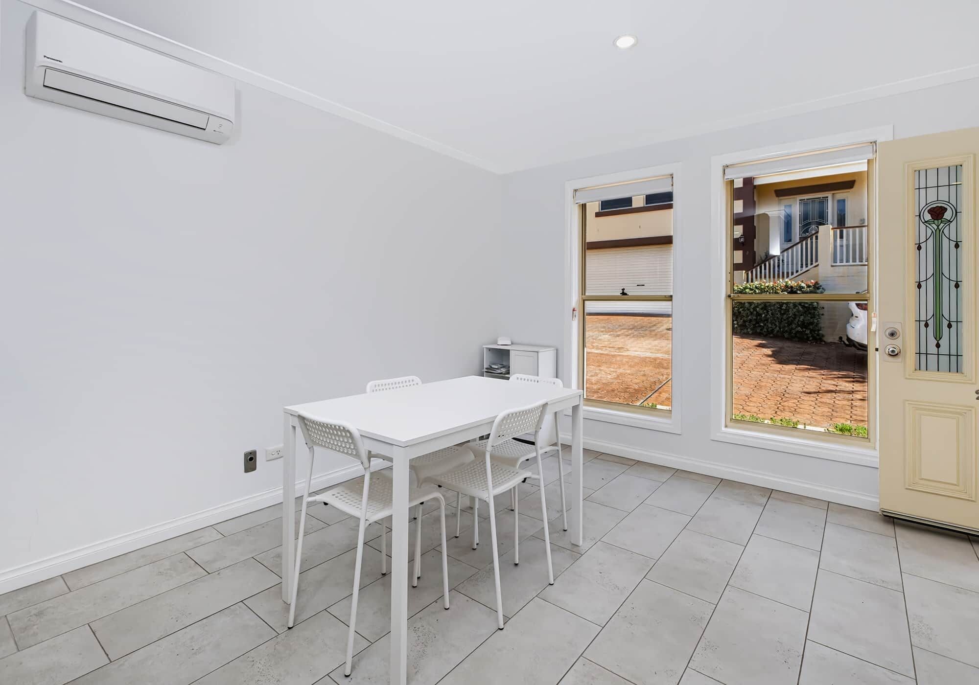 A room with a white table and chairs, a window with a view of a house, and an air conditioning unit on the wall.