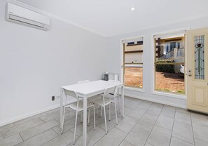 A room with a white table and chairs, a window with a view of a house, and an air conditioning unit on the wall. - Ocean Tracks - 2/1 Eddy Street Kiama (Kiama)