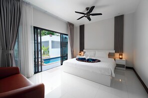 A modern hotel room with a large bed, a ceiling fan, a nightstand with lamps, and a view of a swimming pool. - Elyana Haven | Luxury 3BR Private Pool Villa in Rawai, Phuket (Tambon Rawai)