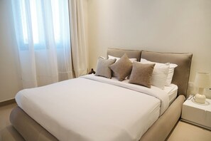 Cama bem arrumada com lençóis brancos e almofadas bege, luminária de cabeceira e janela com cortinas translúcidas.