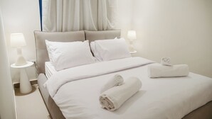 Een keurig opgemaakt bed met witte lakens, twee bedlampen en opgerolde handdoeken.