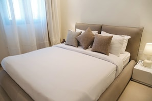 Una cama bien hecha con cabecera, lámpara de noche y ventana con cortinas traslúcidas.