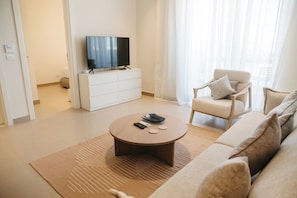 Una sala de estar moderna con un televisor de pantalla plana sobre un mueble blanco, una mesa de centro redonda con controles remotos, un sofá color beige y un sillón con cojines.