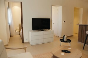 Ein modernes Wohnzimmer mit einem Flachbildfernseher auf einer weißen Kommode, einem runden Couchtisch mit Fernbedienungen und einem Essbereich mit Holztisch und Stühlen.