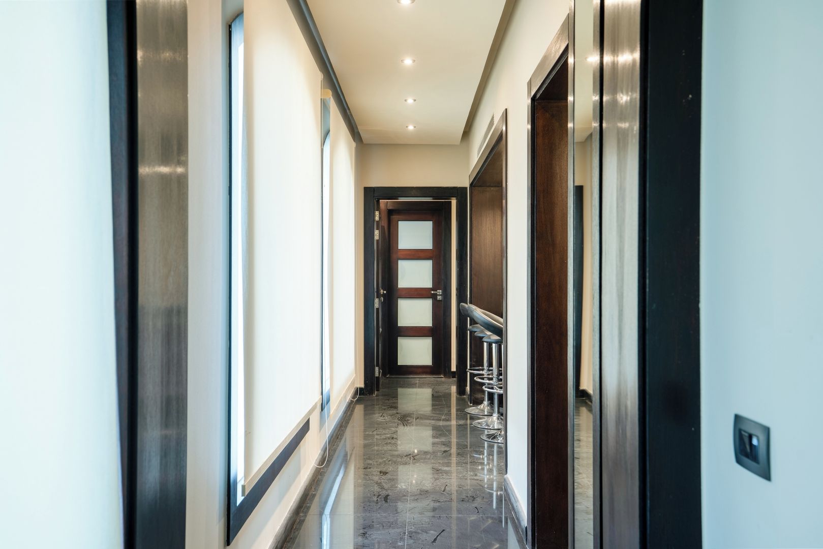 A modern hotel hallway with a marble floor, a wooden door at the end, and a bar area with stools.