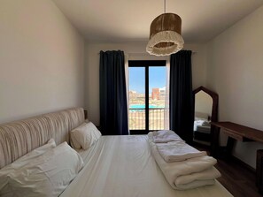 Een hotelkamer met een bed, bureau, spiegel en uitzicht op een zwembad en gebouwen.