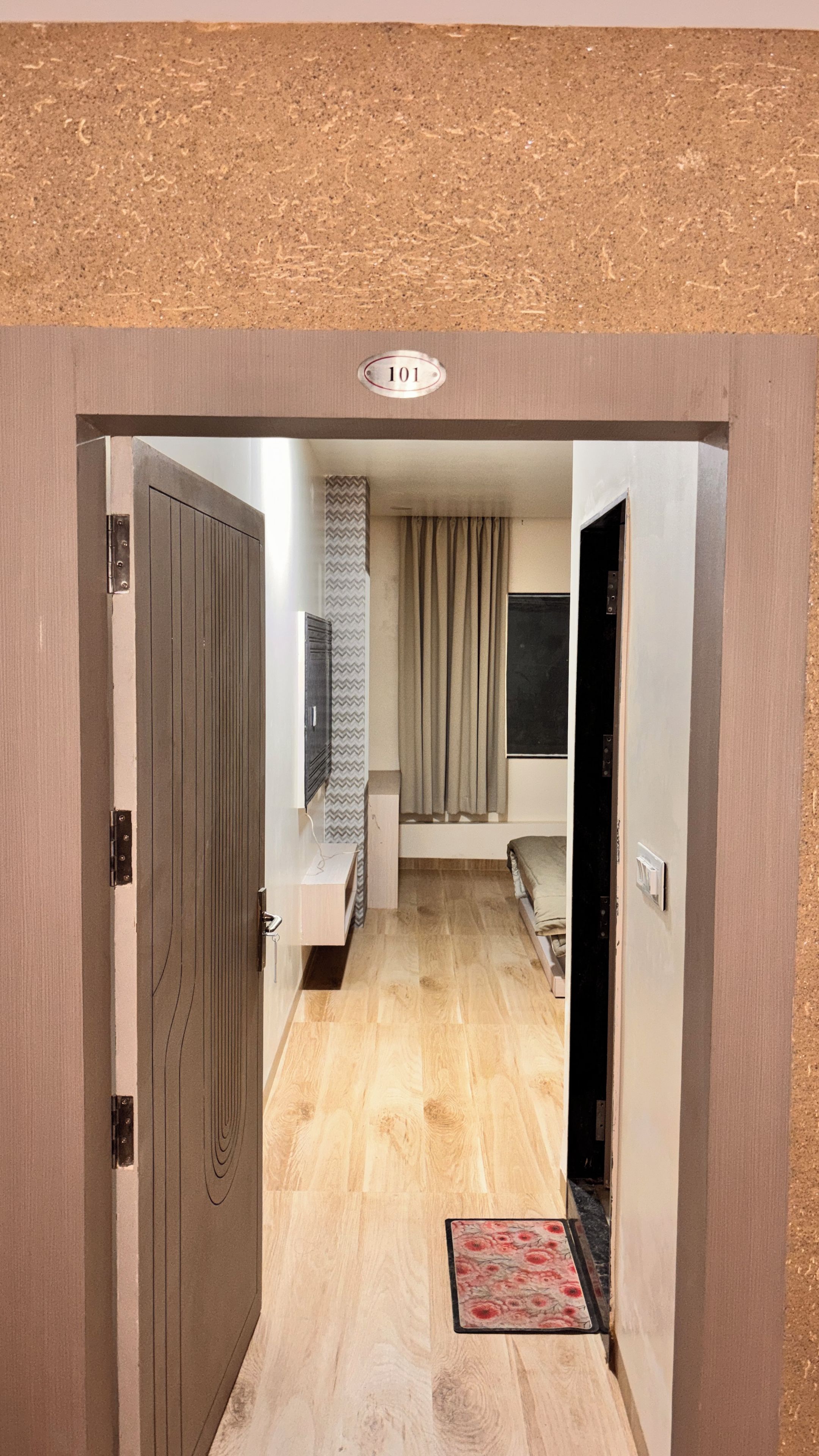 A hallway with a wooden door, a number plate, a bench, a patterned rug, and a bed with a headboard.