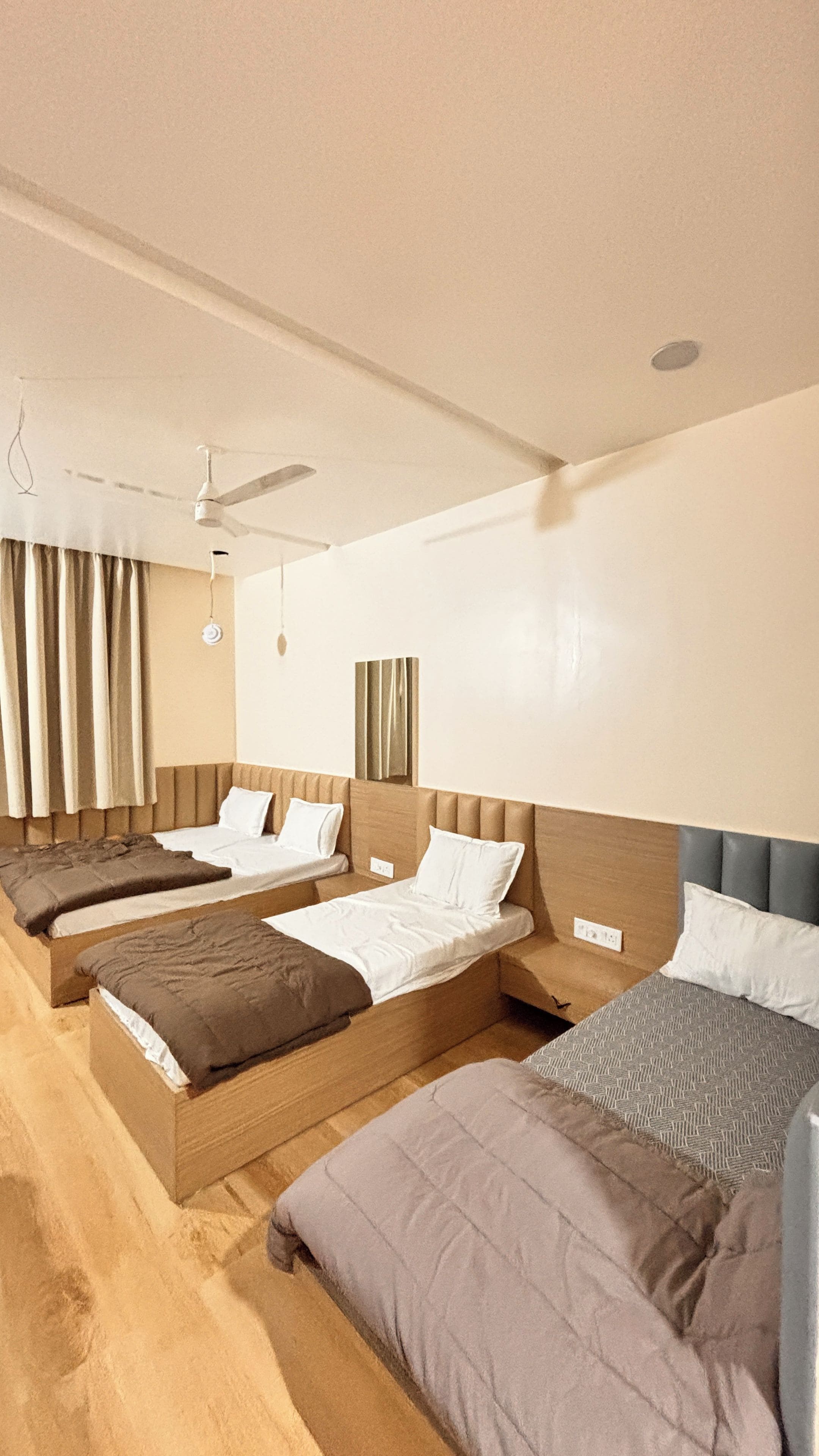 Habitación de hotel con tres camas individuales, un ventilador de techo y una ventana grande con cortinas.