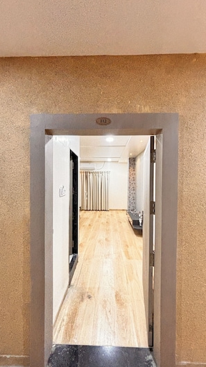A corridor with a wooden floor, a door with a number plate, and a curtain. - The Grand Avantika Hotel and Resorts (Tarana)