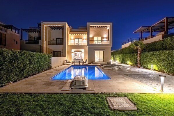 A modern two-story house with a swimming pool, outdoor seating, and well-lit windows.