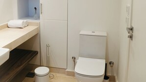 A small, white bathroom with a toilet, a trash can, and a shelf with towels. - 1 BR Ancient Sands Residence (Hurghada)