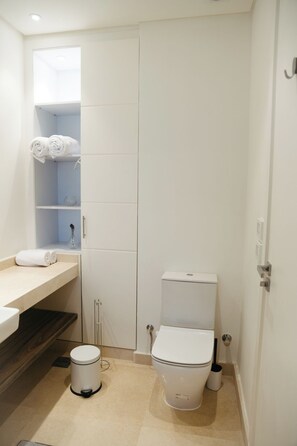 Ein kleines, weißes Badezimmer mit Toilette, einem Mülleimer und einem Ablagefach mit Handtüchern.