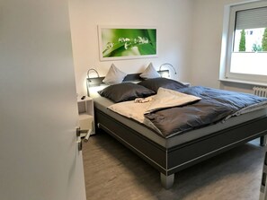 A modern bedroom with a bed, bedside tables, a nightstand, a window with blinds, and a framed picture on the wall. - Holiday Apartment With 1 Bedroom (Goslar)