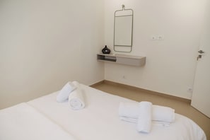 Habitación de hotel con cama, espejo en la pared, estante pequeño con un florero y una puerta.