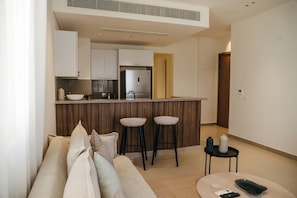 A compact living space with a kitchenette, a sofa with cushions, two bar stools, and a small round table with a remote control. - 1 BR Ancient Sands Residence (Hurghada)