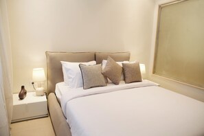 Una cama bien hecha con ropa blanca y varias almohadas, una lámpara de noche y un florero en la mesita de noche.