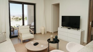 Un salon moderne comprenant un canapé, des fauteuils, une table basse, un téléviseur posé sur un meuble blanc, et une porte-fenêtre coulissante donnant sur un espace extérieur.