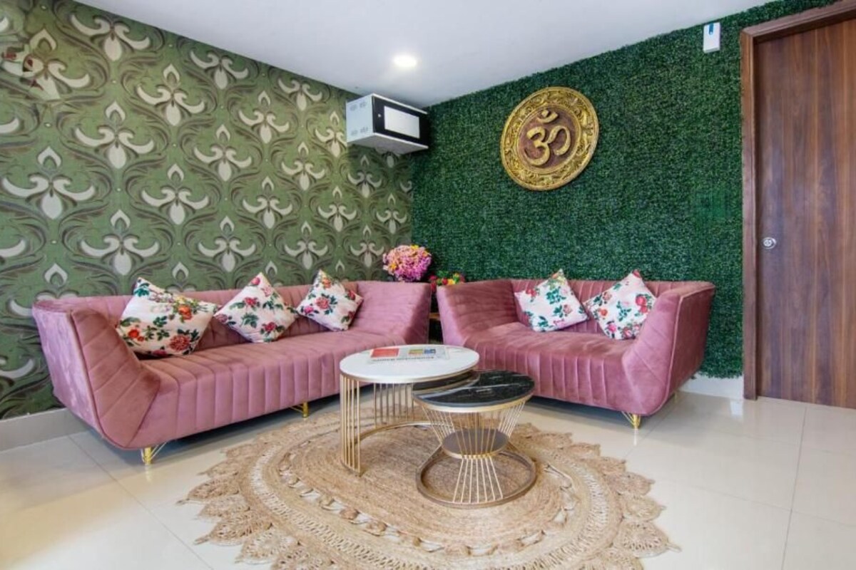 Una habitación con papel tapiz verde estampado, un sofá rosa con cojines florales, una mesa de centro redonda y un aire acondicionado fijado a la pared.